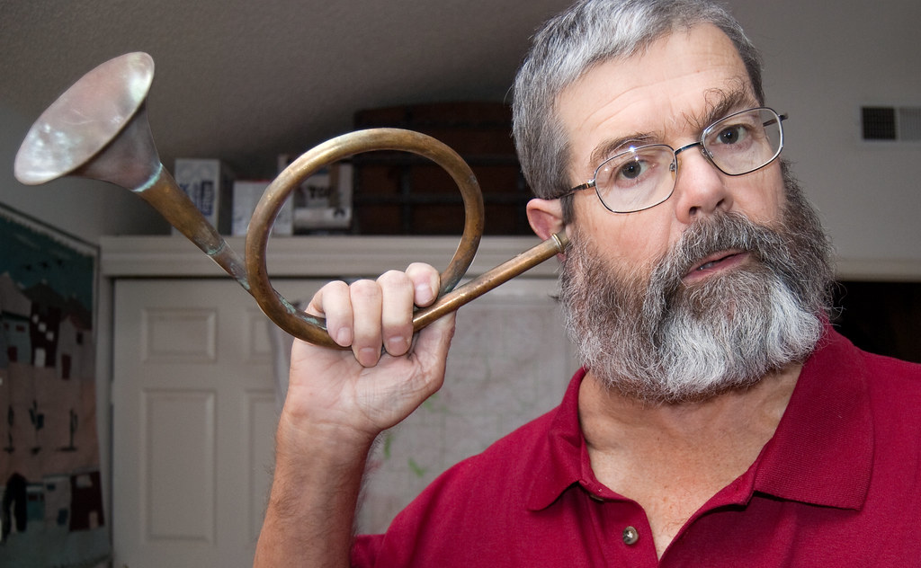 A bearded man holds a coiled ear trumpet to his right ear.